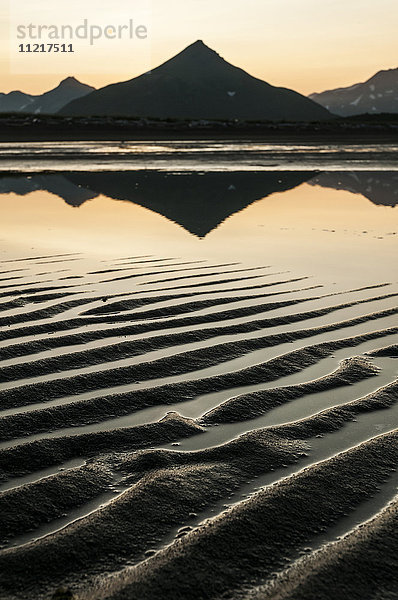 Silhouette eines spitzen Berges  der sich in ruhigem Wasser und Wellen im Sand bei Sonnenuntergang spiegelt; Alaska  Vereinigte Staaten von Amerika'.