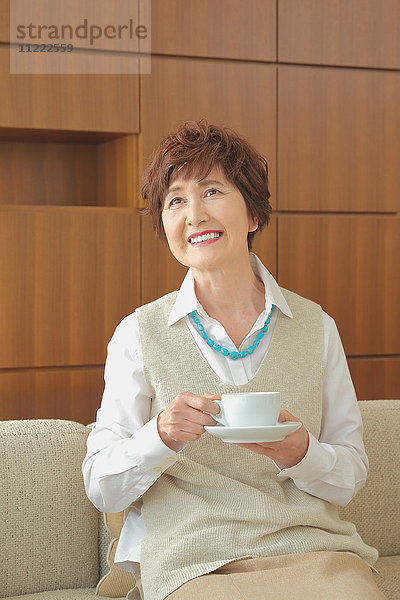 Modische japanische Seniorin  die auf dem Sofa eine Tasse Kaffee trinkt
