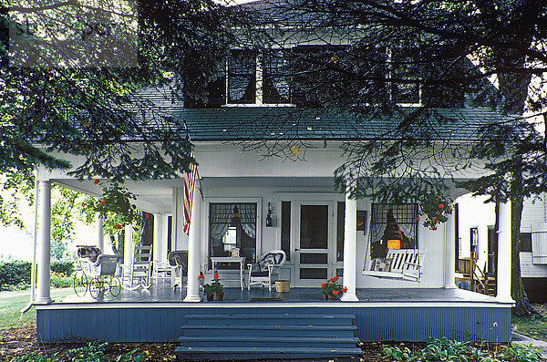 Hübsches Sommerhaus mit großer Veranda mit Säulen. Veranda rundherum. Gaubenfenster. Amerikanische Flagge  Weidenwagen.