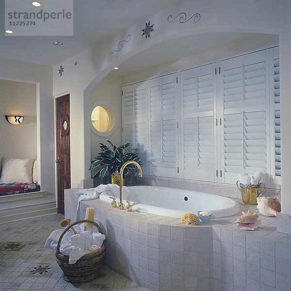 Badewanne mit Flieseneinfassung in gewölbter Nische mit Fensterläden  rundes Fenster. Korb mit Handtüchern auf gefliestem Boden  südwestliche Muster  rundes Fenster