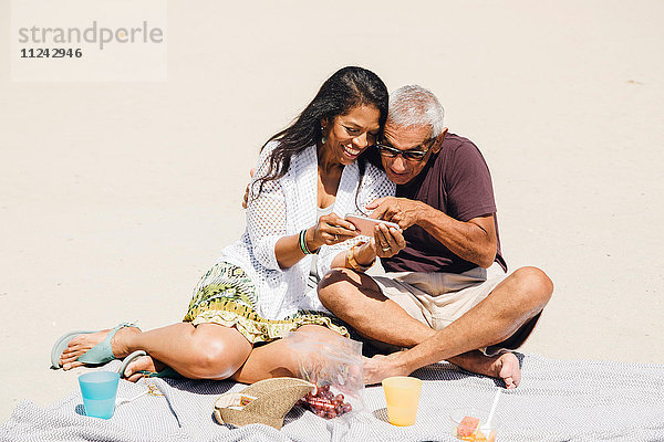 Älteres Ehepaar sitzt beim Picknick am Strand und schaut sich ein Smartphone an  Long Beach  Kalifornien  USA