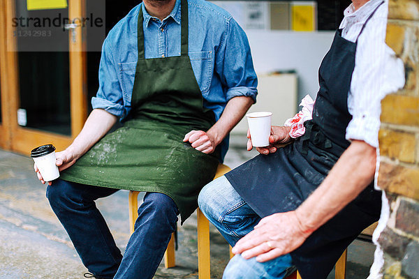 Älterer Handwerker trinkt Kaffee und unterhält sich mit einem jungen Mann außerhalb der Werkstatt  mittlerer Abschnitt