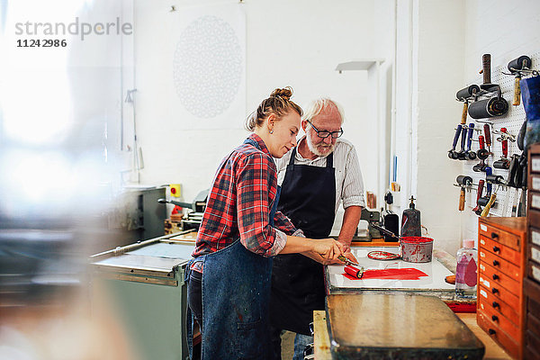Leitender Handwerker/Techniker beaufsichtigt junge Frau  wie sie in einer Buchkunstwerkstatt Farbwalze für den Buchdruck verwendet