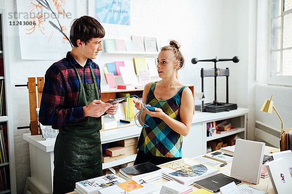 Junge Frau unterhält sich mit einem jungen männlichen Handwerker und nutzt die Tablett-Technologie  um im Kunstbuchladen einzukaufen