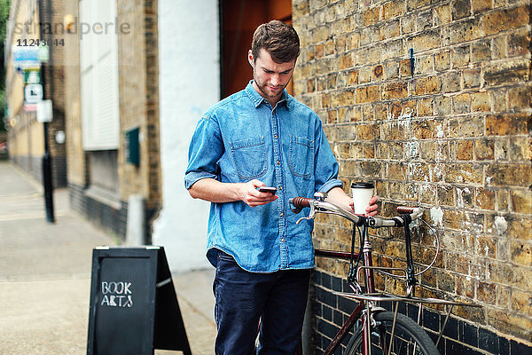 Junger Mann hält Kaffee in der Hand und überprüft Handy  mit an Ziegelmauer gelehntem Schubfahrrad
