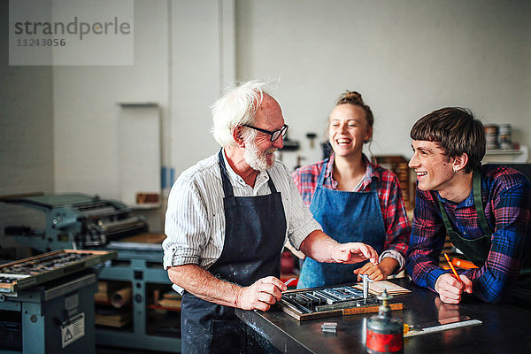 Älterer Handwerker lacht mit jungem Handwerker und Handwerkerin in Buchdruckwerkstatt