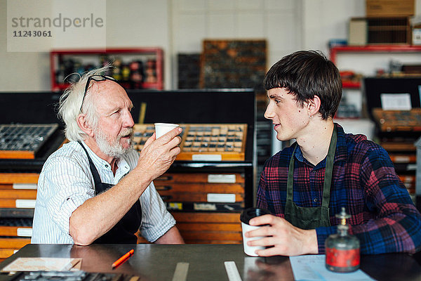 Älterer Handwerker trinkt Kaffee und unterhält sich mit einem jungen Handwerker in einem Buchkunst-Workshop