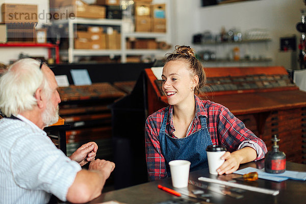 Junge Handwerkerin trinkt Kaffee  plaudert und lacht mit einem älteren Handwerker in einem Buchkunst-Workshop