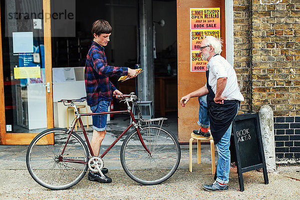 Junger Mann mit Fahrrad schaut auf Posten und steht mit einem älteren Handwerker vor der Druckerei
