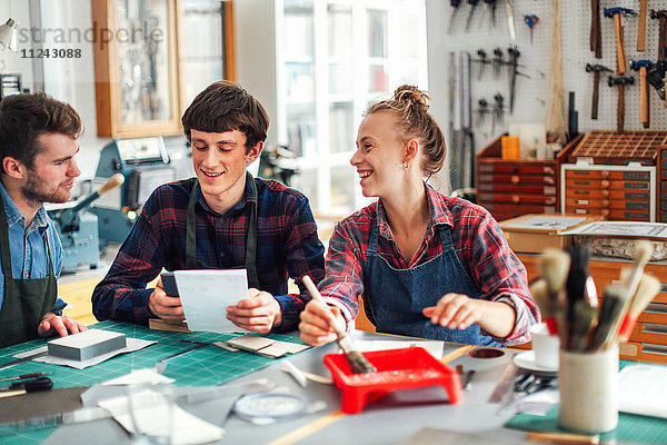 Junge Handwerkerin hält Pinsel und lacht und lächelt mit zwei jungen Handwerkern im kreativen Druckatelier