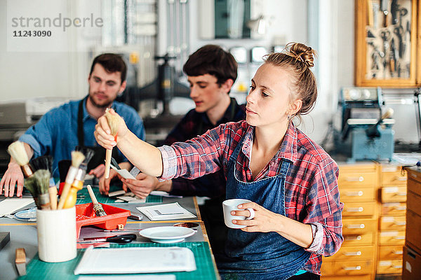 Junge Handwerkerin hält Kaffeetasse und greift nach dem Pinsel im kreativen Druckatelier  im Hintergrund arbeiten zwei junge Handwerker