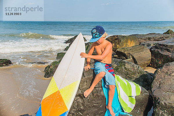 Junge sitzt am Strand auf Felsen und hält ein Surfbrett