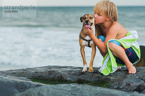 Kleiner Junge kauert am Strand auf Felsen und umarmt Haushund