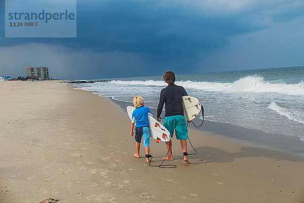 Vater und Sohn gehen am Strand entlang  tragen Surfbretter  Rückansicht