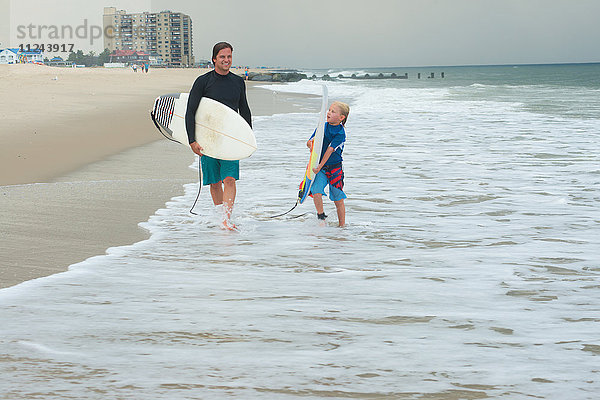 Vater und Sohn gehen am Strand spazieren und tragen Surfbretter