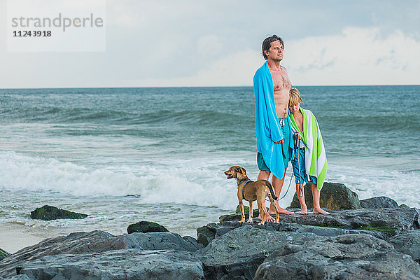 Vater und Sohn stehen auf Felsen am Meer  Strandtücher um sie herum drapiert  daneben ein Hund als Haustier