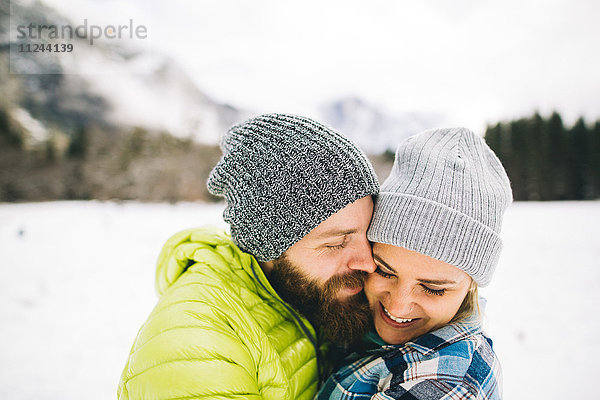 Paar auf schneebedeckter Landschaft mit Strickmützen umarmend