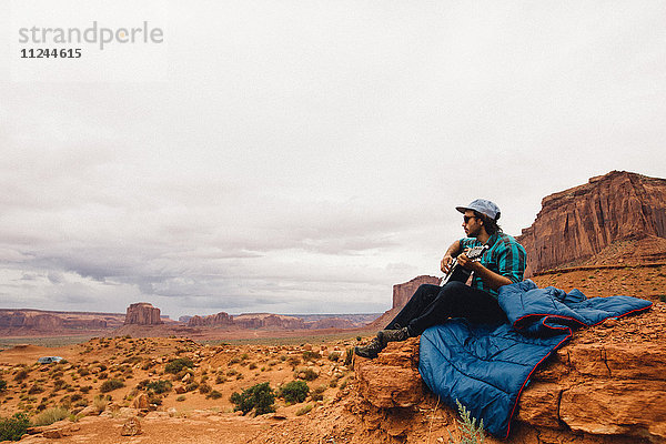 Auf Rock sitzender junger Mann spielt Akustikgitarre  Monument Valley  Arizona  USA