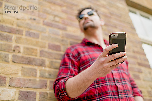 Niedrigwinkelansicht eines jungen Mannes  der mit einem Smartphone an einer Ziegelmauer lehnt