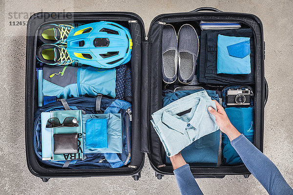 Draufsicht auf die Hände eines Mannes  der einen Koffer mit Wanderschuhen  Fahrradhelm  Rucksack  Retro-Kamera und blauem Hemd packt