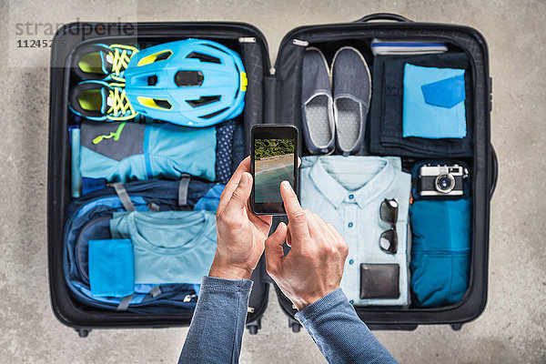 Draufsicht auf die Hände des Mannes mittels Smartphone-Touchscreen über gepacktem Koffer mit blauem Fahrradhelm  Rucksack  Retro-Kamera und Hemd