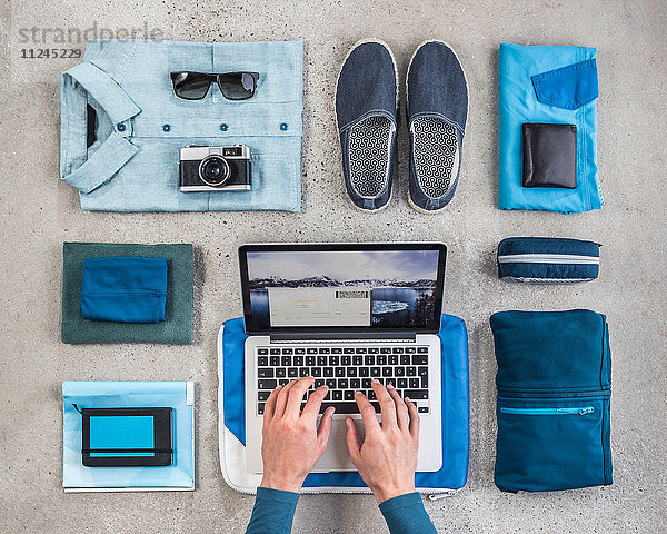 Draufsicht auf die Hände eines Mannes beim Tippen am Laptop  umgeben von Reisepackstücken  mit blauem Hemd  Retro-Kamera  Waschbeutel und Notizbuch