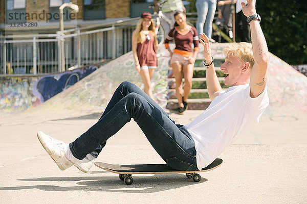 Junger männlicher Skateboardfahrer sitzt auf einem Skateboard und bewegt sich im städtischen Skatepark