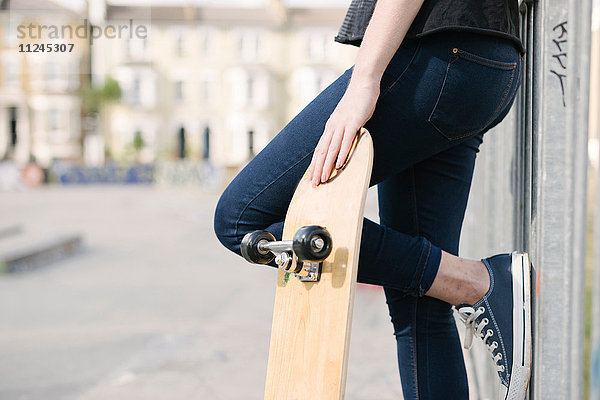 Taille einer Skateboardfahrerin  die sich im Skatepark gegen einen Zaun lehnt