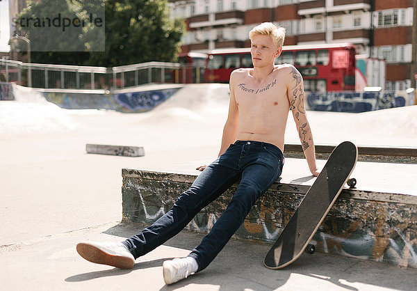 Tätowierter junger männlicher Skateboarder sitzt im Skatepark