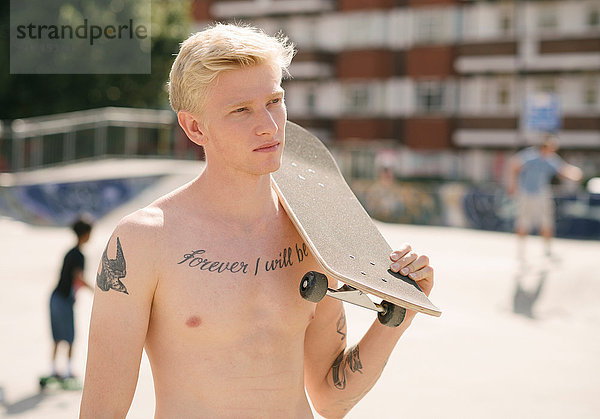 Tätowierter junger Mann mit Skateboard auf der Schulter im Skatepark