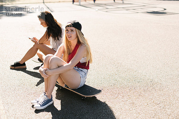 Zwei erwachsene Freundinnen sitzen auf Skateboards auf dem Basketballplatz