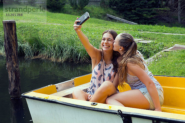 Zwei junge Freundinnen beim Smartphone-Selfie im Ruderboot am Seeufer