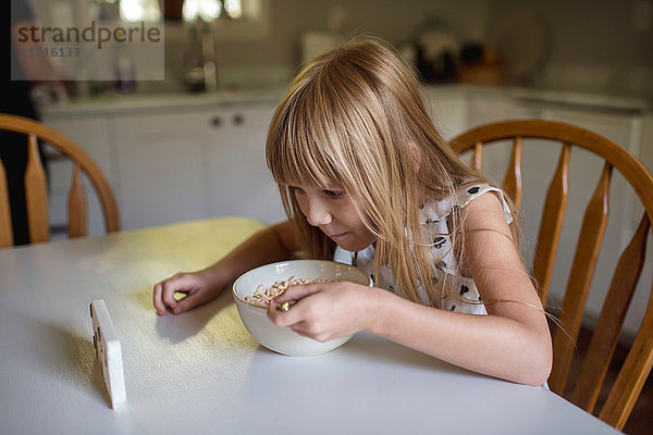 Kleines Mädchen beim Frühstücken  während es sich ein elektronisches Gerät ansieht.