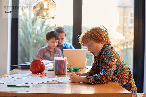 Junge malt am Esstisch  während der Vater mit seinem Bruder am Laptop sitzt