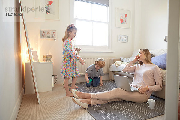 Mittlere erwachsene Frau sitzt mit Laptop auf dem Wohnzimmerboden  während Sohn und Tochter spielen