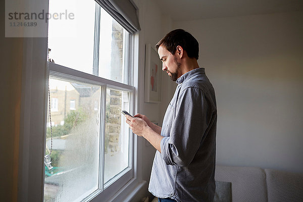 Mittlerer Erwachsener Mann am Wohnzimmerfenster beim Lesen von Smartphone-Texten