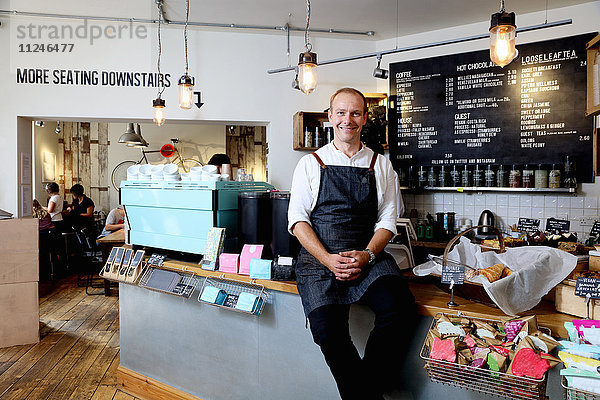 Porträt eines männlichen Geschäftsinhabers  der auf dem Tresen eines unabhängigen Cafés sitzt