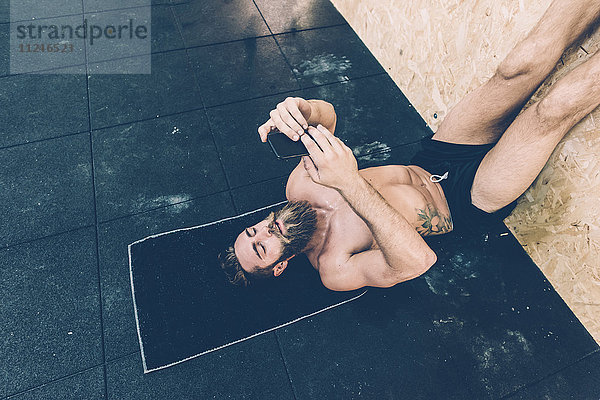 Männlicher Crosstrainer liest im Fitnessstudio auf dem Rücken liegend ein Smartphone