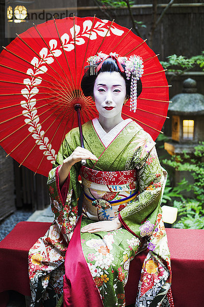 Eine im traditionellen Geisha-Stil gekleidete Frau in Kimono und Obi  mit aufwändiger Frisur und blumigen Haarspangen  mit weißer Gesichtsschminke mit leuchtend roten Lippen und dunklen Augen  die einen Regenschirm hält.
