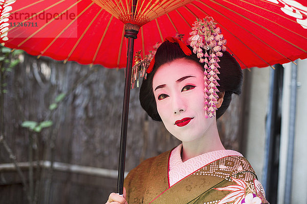 Eine im traditionellen Geisha-Stil gekleidete Frau  in einem Kimono mit aufwändiger Frisur und blumigen Haarspangen  mit weißer Gesichtsschminke mit leuchtend roten Lippen und dunklen Augen  die einen roten Papiersonnenschirm halten.