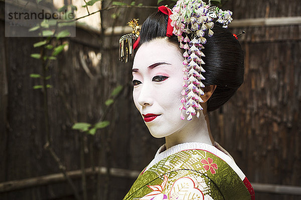 Eine im traditionellen Geisha-Stil gekleidete Frau in Kimono und Obi  mit aufwändiger Frisur und blumigen Haarspangen  mit weißer Gesichtsschminke mit leuchtend roten Lippen und dunklen Augen.
