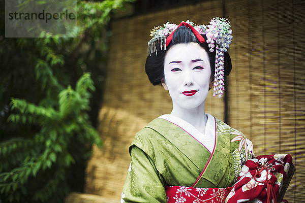 Eine im traditionellen Geisha-Stil gekleidete Frau in Kimono und Obi  mit aufwändiger Frisur und blumigen Haarspangen  mit weißer Gesichtsschminke mit leuchtend roten Lippen und dunklen Augen.