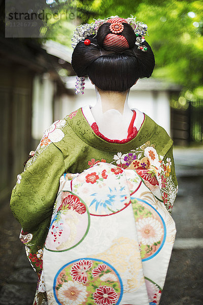 Eine im traditionellen Geisha-Stil gekleidete Frau in Kimono und Obi  mit aufwändiger Frisur und blumigen Haarspangen  mit weißer Gesichtsschminke mit leuchtend roten Lippen und dunklen Augen auf einer Straße.