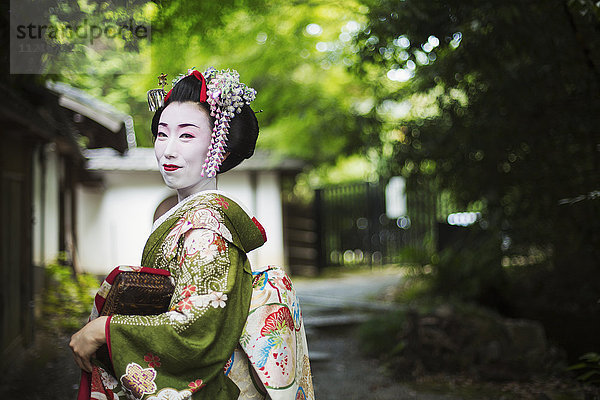 Eine im traditionellen Geisha-Stil gekleidete Frau in Kimono und Obi  mit aufwändiger Frisur und blumigen Haarspangen  mit weißer Gesichtsschminke mit leuchtend roten Lippen und dunklen Augen  Seitenansicht im Freien.