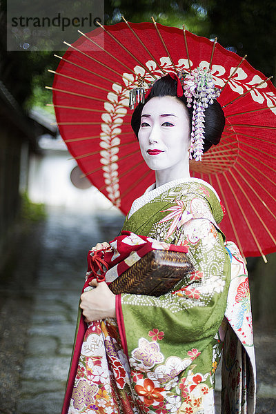 Eine im traditionellen Geisha-Stil gekleidete Frau  in Kimono und Obi  mit aufwändiger Frisur und blumigen Haarspangen  mit weißer Gesichtsschminke mit leuchtend roten Lippen und dunklen Augen  die einen roten Papiersonnenschirm halten.