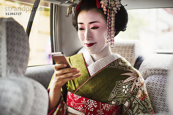 Eine im traditionellen Geisha-Stil gekleidete Frau in Kimono und Obi  mit aufwändiger Frisur und blumigen Haarspangen  mit weißem Gesichts-Make-up mit leuchtend roten Lippen und dunklen Augen in einem Auto mit einem Smartphone.