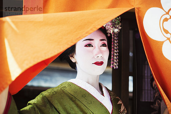 Eine im traditionellen Geisha-Stil gekleidete Frau  in einem Kimono mit aufwändiger Frisur und blumigen Haarspangen  mit weißer Gesichtsschminke mit leuchtend roten Lippen und dunklen Augen  die einen orangefarbenen Vorhang hochziehen.