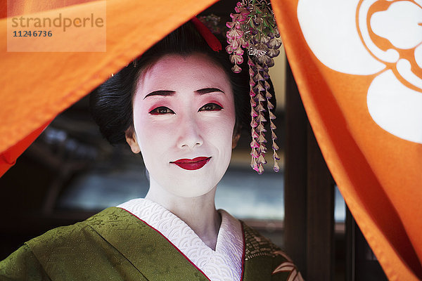 Eine im traditionellen Geisha-Stil gekleidete Frau  in einem Kimono mit aufwändiger Frisur und blumigen Haarspangen  mit weißer Gesichtsschminke mit leuchtend roten Lippen und dunklen Augen  die einen orangefarbenen Vorhang hochziehen.