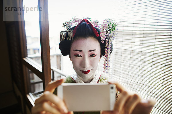 Eine im traditionellen Geisha-Stil gekleidete Frau  in Kimono und Obi  mit aufwändiger Frisur und blumigen Haarspangen  mit weißer Gesichtsschminke  mit leuchtend roten Lippen und dunklen Augen  die ein Selfie von sich gibt.