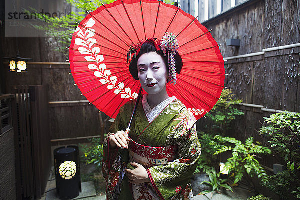 Eine im traditionellen Geisha-Stil gekleidete Frau  in Kimono und Obi  mit aufwändiger Frisur und blumigen Haarspangen  mit weißer Gesichtsschminke mit leuchtend roten Lippen und dunklen Augen  die einen roten Papiersonnenschirm halten.
